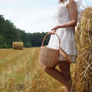 May include: A woman in a white dress holds a wicker basket in a field of hay bales. The basket has a light brown color and a lid.