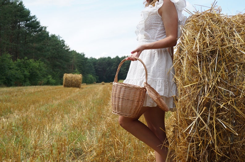 May include: A woman in a white dress holds a wicker basket in a field of hay bales. The basket has a light brown color and a lid.