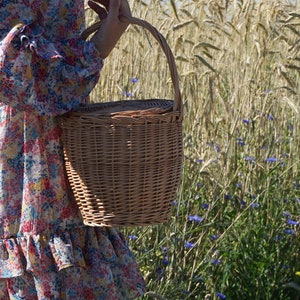 May include: A woman in a floral dress holds a wicker basket with a handle. The basket is brown and has a woven texture. The woman is standing in a field of tall grass and blue flowers.