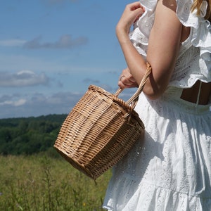 May include: A woman wearing a white dress with lace detailing carries a woven wicker basket with a handle. The basket is brown and has a natural woven texture. The woman is standing in a field with green grass and a blue sky in the background.