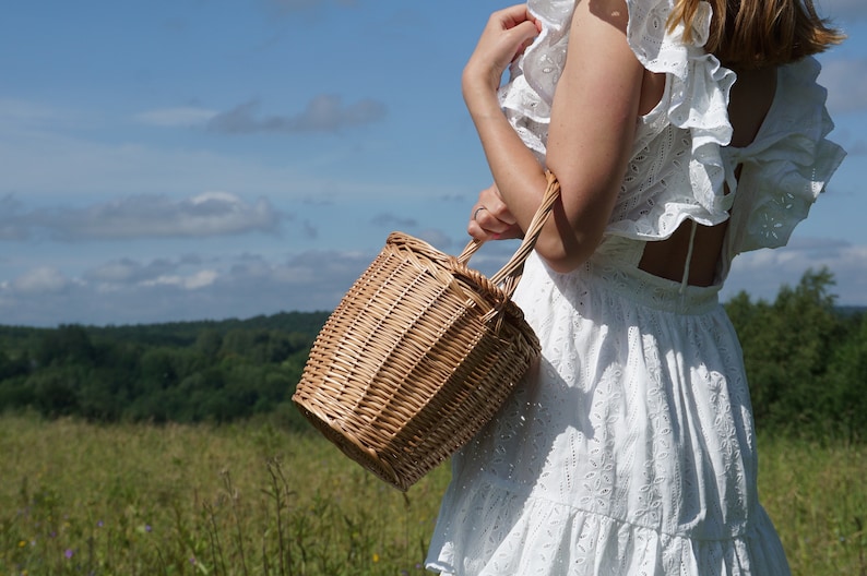 May include: A woman wearing a white dress with lace detailing carries a woven wicker basket with a handle. The basket is brown and has a natural woven texture. The woman is standing in a field with green grass and a blue sky in the background.