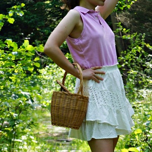 May include: A woman wearing a pink sleeveless shirt and a white eyelet skirt with a ruffled hem. She is holding a woven wicker basket.