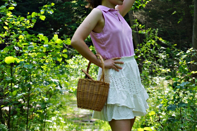 May include: A woman wearing a pink sleeveless shirt and a white eyelet skirt with a ruffled hem. She is holding a woven wicker basket.