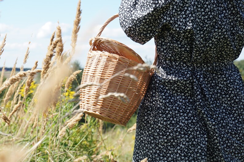May include: A woman in a black and white floral dress carries a woven wicker basket with a handle.