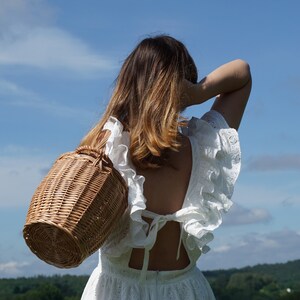 May include: A woman wearing a white dress with a lace overlay and a wicker basket slung over her shoulder. The dress has a low back with a tie detail. The woman is standing in a field with a blue sky and white clouds in the background.