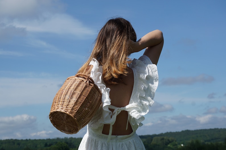 May include: A woman wearing a white dress with a lace overlay and a wicker basket slung over her shoulder. The dress has a low back with a tie detail. The woman is standing in a field with a blue sky and white clouds in the background.