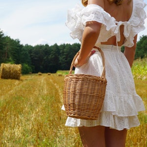 May include: A woman in a white, ruffled dress holds a wicker basket in a field of golden hay.