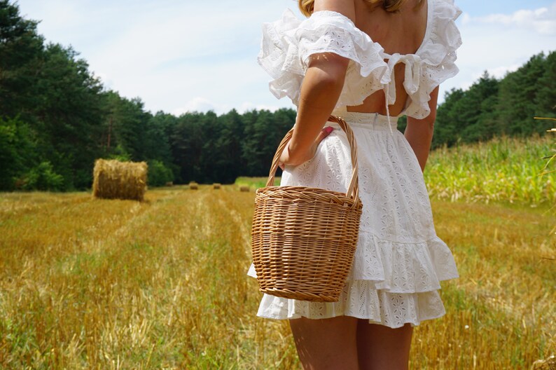 May include: A woman in a white, ruffled dress holds a wicker basket in a field of golden hay.