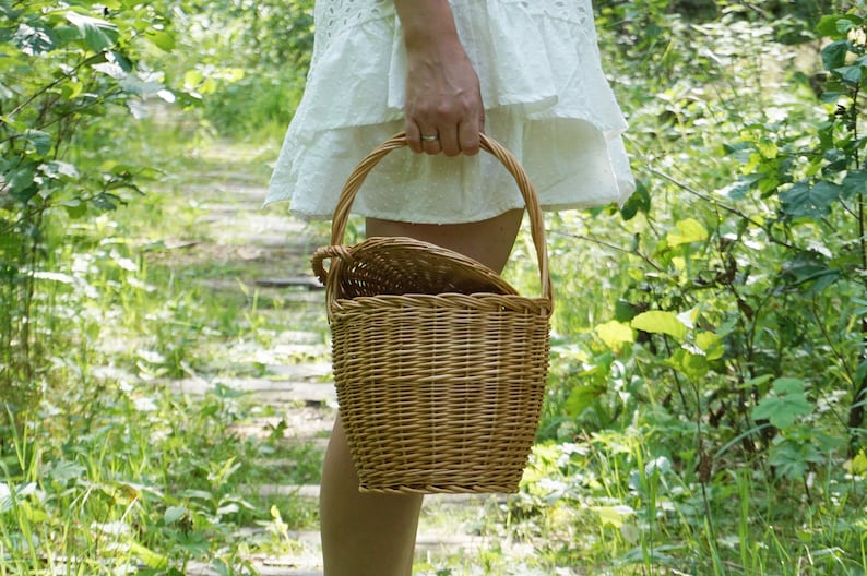 May include: A woman in a white dress carries a brown wicker basket with a handle. The basket is woven with a natural, light brown material. The basket is in a forest setting with green foliage.