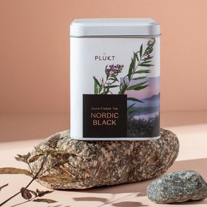 Puede incluir: Una lata blanca de té con una etiqueta que dice "PLUKT Hand Picked Tea NORDIC BLACK". La etiqueta presenta una fotografía de una planta con flores moradas y un paisaje brumoso.
