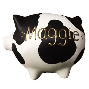 May include: Black and white cow-print piggy bank with the name "Maggie" written in gold lettering.