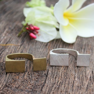 May include: Two open-ended rings, one gold and one silver, on a wooden surface. The rings are both rectangular in shape and have a brushed finish.