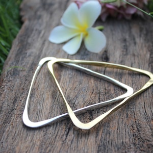 May include: Two triangular bangles, one silver and one gold, rest on a weathered wooden surface. A white and yellow flower sits above the bangles, with green grass in the background. The bangles are a simple, geometric design.