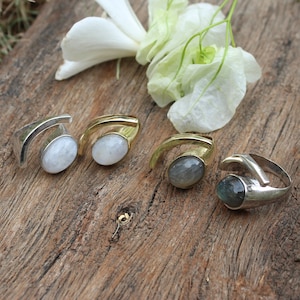 May include: Three silver and gold rings with different colored gemstones. The rings are on a wooden surface with white flowers.
