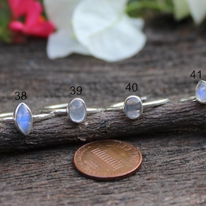 May include: A collection of silver rings with various gemstone shapes, including round, oval, and teardrop. The rings are displayed on a dark wooden branch, with a penny for size comparison. The gemstones have a blue and white iridescent effect.
