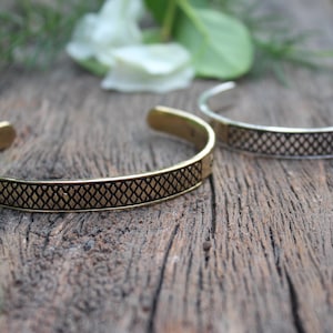 May include: Two open cuff bracelets. One is gold-toned, the other silver-toned. Both feature a repeating diamond pattern. The bracelets are on a weathered wooden surface, with greenery and a white flower in the background.