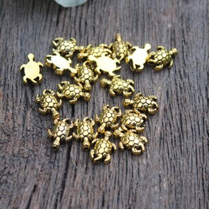 May include: A group of 20 gold-colored turtle charms, scattered on a wooden surface. The charms are detailed with a textured shell design.