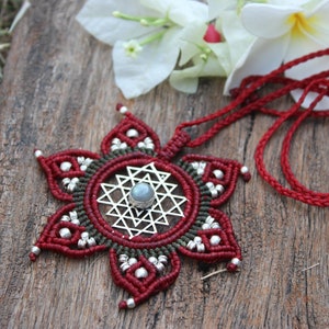 Puede incluir: Un collar con un colgante de macramé en forma de flor roja y verde con un amuleto Sri Yantra plateado y una piedra de luna en el centro. El collar está sobre una superficie de madera con flores blancas.