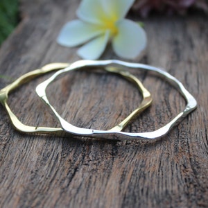 Può includere: Due semplici braccialetti, uno d'argento e uno d'oro, con una forma leggermente irregolare e ondulata. I braccialetti sono appoggiati su un pezzo di legno invecchiato.