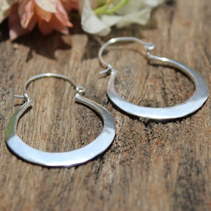 May include: A pair of silver hoop earrings with a slightly curved, flat design. The earrings are on a wooden surface.