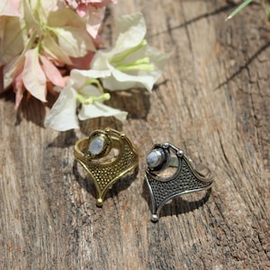 May include: Two silver rings with moonstone settings. The rings have an intricate design and are set on a wooden surface.