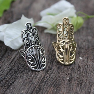 May include: Two hamsa hand rings, one silver and one gold, with intricate filigree designs. The rings are on a wooden surface.
