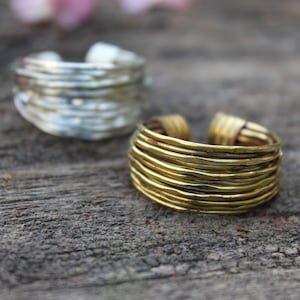 Puede incluir: Dos anillos de color dorado con un diseño en capas y martillado. Los anillos están apilados uno encima del otro sobre una superficie de madera.