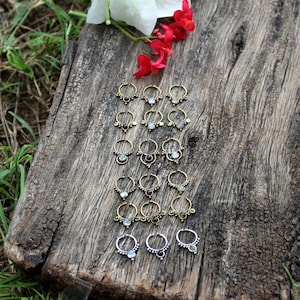 Puede incluir: Una colección de 20 anillos de nariz dorados con piedras preciosas de varios colores. Los anillos están dispuestos en filas sobre una superficie de madera.