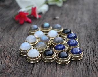 May include: A collection of gemstone cabochons set in gold-toned bezels. The stones include moonstone, labradorite, and lapis lazuli. The cabochons are arranged in a triangular pattern on a wooden surface.