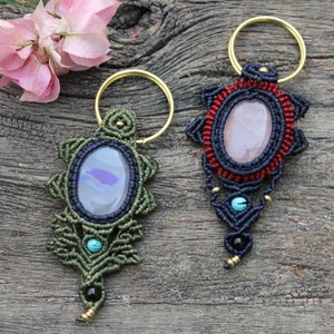May include: Two macrame keychains with gemstone accents. The keychain on the left has a green and blue macrame design with a light blue agate stone. The keychain on the right has a red, blue, and black macrame design with a pink rose quartz stone.