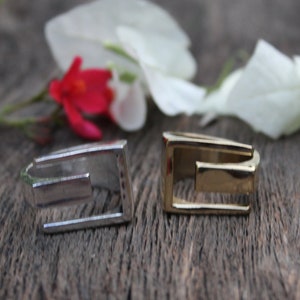 Puede incluir: Dos anillos de metal, uno plateado y otro dorado, con forma cuadrada y un diseño recortado. Los anillos están sobre una superficie de madera.