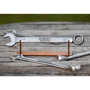 Peut inclure: Une clé à molette argentée gravée du nom "ROBERT" et du mot "GROOMSMAN". La clé est exposée sur un support en bois. D'autres clés sont visibles en arrière-plan.