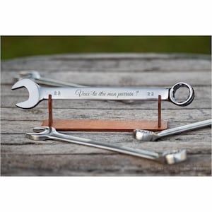 Könnte beinhalten: Ein silberner Schraubenschlüssel, Größe 22, mit dem französischen Text "Veux-tu être mon parrain ?" graviert, auf einem Holzständer präsentiert. Weitere Schraubenschlüssel sind auf einer Holzoberfläche im Hintergrund zu sehen.