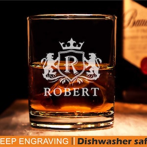 May include: A clear glass with a deep engraving of a crest featuring a lion, a crown, and the letter "R" with the name "ROBERT" below. The glass is filled with amber liquid.