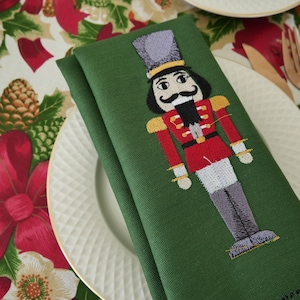 May include: A folded green cloth napkin with an embroidered nutcracker design. The nutcracker is depicted in a red jacket, gray pants, and a tall hat. The napkin is placed on a white plate with a gold rim, with a festive floral tablecloth in the background.
