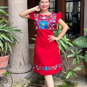May include: A red embroidered dress with colourful floral patterns. The dress has short sleeves and a fitted bodice. The skirt is slightly flared and falls to the knees. The dress is worn by a person standing in front of a wooden door.