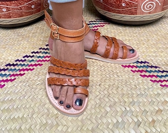 Può includere: Un paio di sandali in pelle marrone con cinturini intrecciati. I sandali hanno una chiusura a fibbia e sono indossati ai piedi.