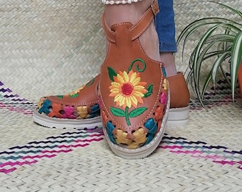 May include: Brown leather sandals with colorful embroidered flowers. The sandals have a strap around the ankle and a woven sole.