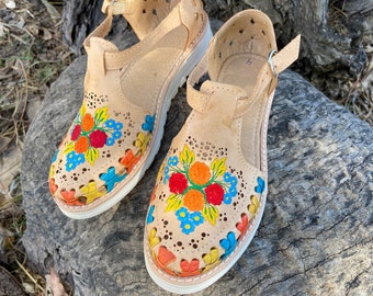 Puede incluir: Un par de sandalias de cuero marrón con bordados florales de colores. Las sandalias tienen una correa sobre el pie y una correa de tobillo con hebilla. Las sandalias tienen una suela blanca.