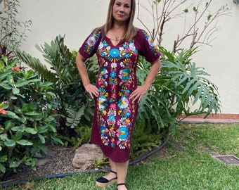 May include: A burgundy embroidered dress with short sleeves and a knee-length hem. The dress features a vibrant floral design in shades of blue, yellow, pink, and white. The woman is wearing black platform sandals. The background includes green plants and grass.