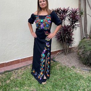 May include: A black maxi dress with colourful floral embroidery. The dress has a square neckline and short sleeves. The embroidery features flowers, butterflies, and leaves in shades of pink, purple, blue, green, orange, and yellow.