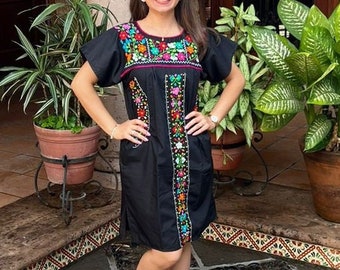 May include: A black embroidered dress with colorful floral patterns and a white headband. The dress has short sleeves and a fitted silhouette. The person is wearing brown leather sandals.