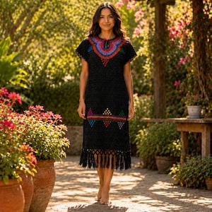May include: A black embroidered dress with colorful geometric patterns around the neckline, waist, and hem. The dress features short sleeves and a fringe hemline. The woman is standing barefoot on a stone path in a garden setting.