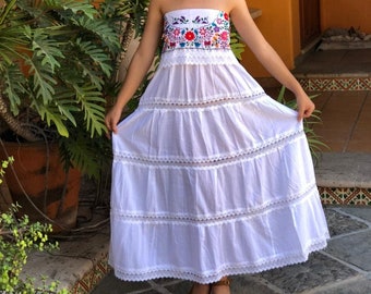 May include: A white strapless maxi dress featuring a vibrant floral embroidery pattern across the bodice, accented with horizontal bands of intricate white lace trim throughout the tiered skirt, paired with a straw hat and brown platform sandals.