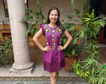 May include: A purple sleeveless dress featuring a square neckline with vibrant floral embroidery in yellow, orange, blue, and green. The bodice includes vertical pleated details, and the hem is finished with a wide, dark purple lace trim.