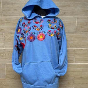 May include: A light blue hooded sweatshirt with colorful embroidered flowers on the front. The sweatshirt has a kangaroo pocket.