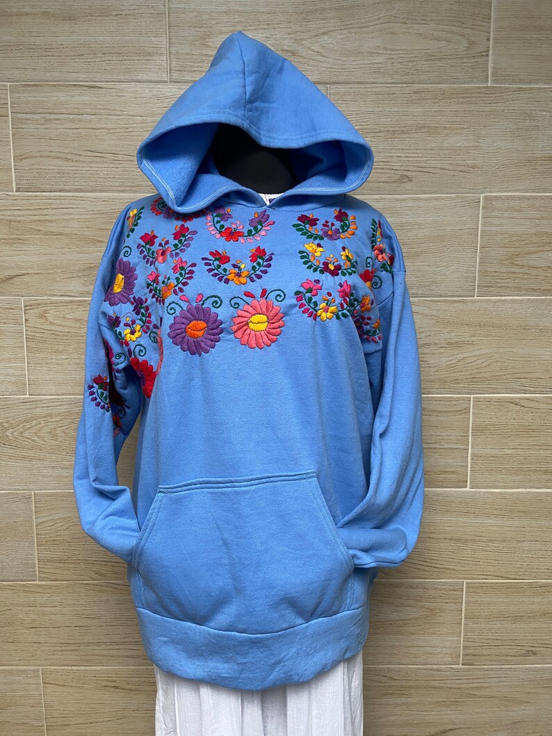 May include: A light blue hooded sweatshirt with colorful embroidered flowers on the front. The sweatshirt has a kangaroo pocket.