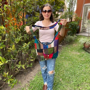 May include: A colorful patchwork tote bag with a brown strap. The bag has a variety of colors and patterns, including red, green, blue, and yellow. The bag is being held by a person wearing a pink shirt and blue jeans.