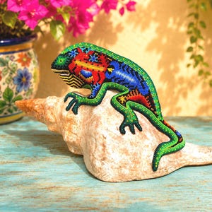 May include: A decorative lizard figurine covered in intricate, multi-colored beadwork featuring vibrant green, blue, red, and yellow patterns. The lizard is positioned on top of a large, textured, off-white conch shell, showcasing detailed craftsmanship.