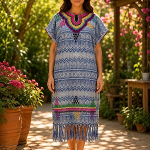 May include: A light blue, knee-length dress with short sleeves and a decorative neckline. The dress features a geometric pattern and colorful embroidery in pink, yellow, and green. The hemline has a fringe detail. The dress is worn outdoors.